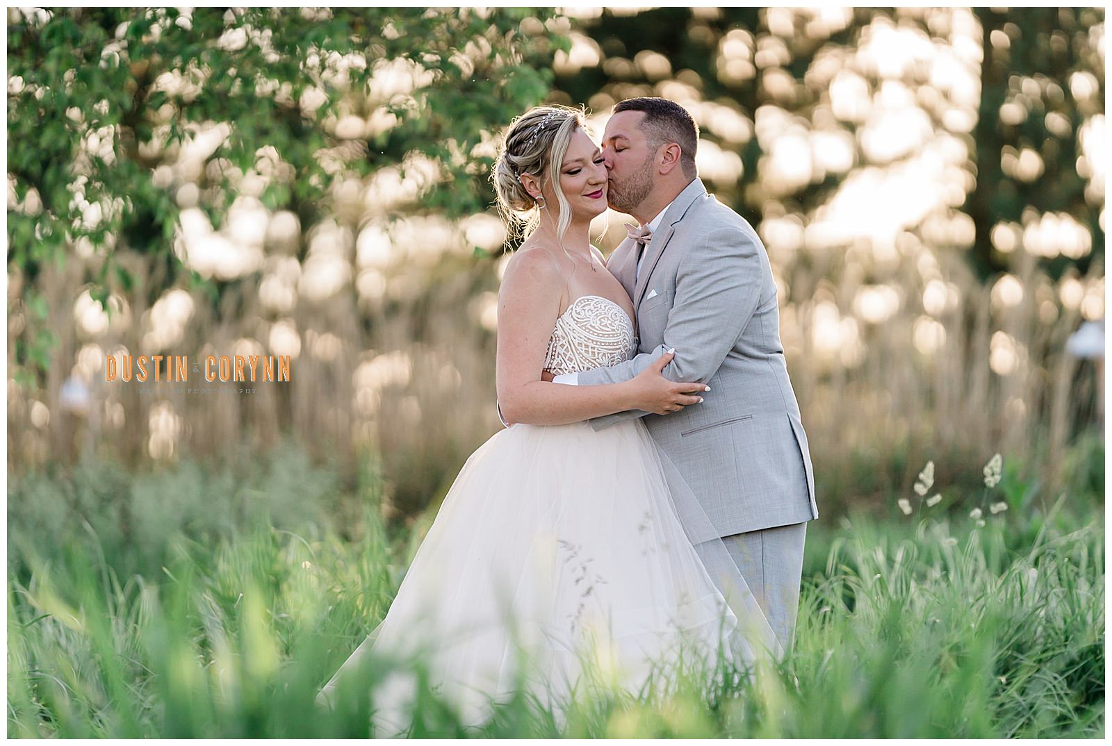 groom holding his bride and kissing her cheek as they stand together in a green grass field in Fort Wayne