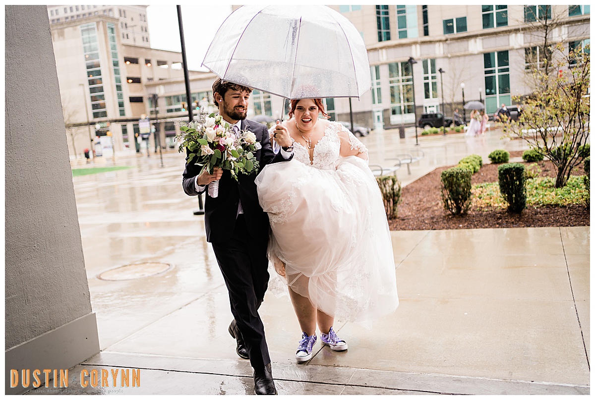 bride and groom under a clear umbrella on their wedding day as they run inside of their Fort Wayne wedding venue