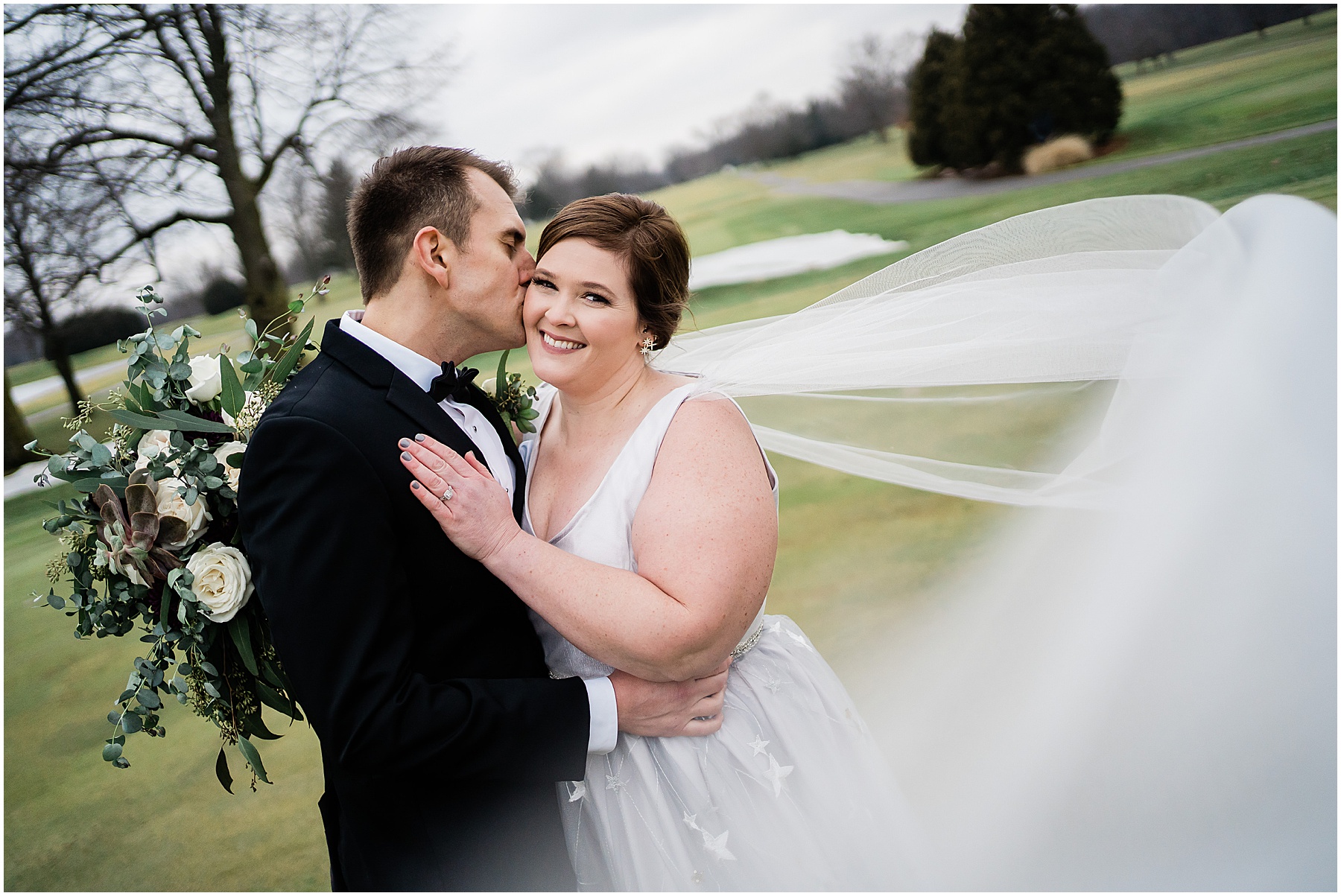 groom kissing his bride on the cheek as she smiles at the camera while her veil blows in the wind at Fort Wayne wedding venue