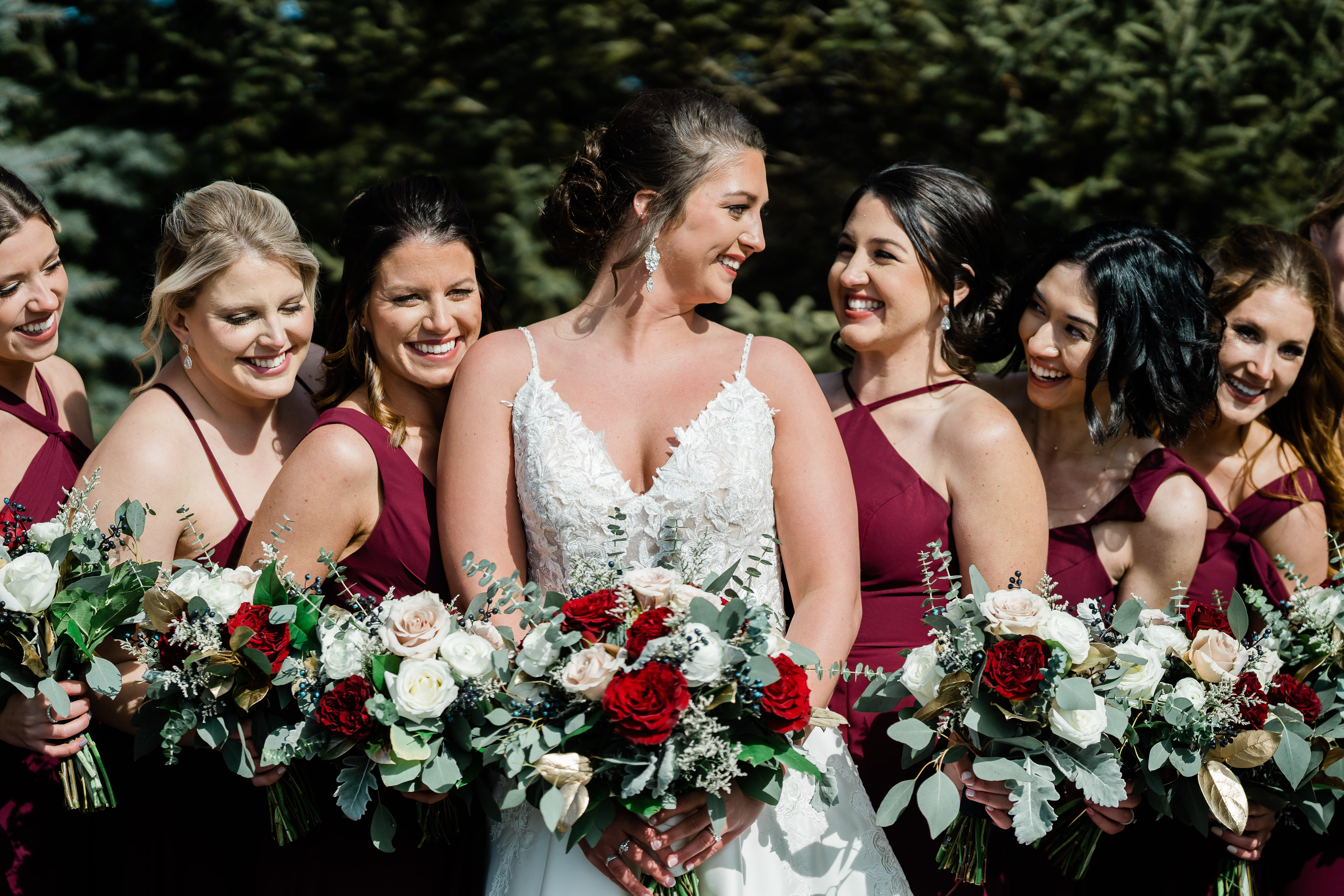 bridesmaids in dark red gown surround the bride as they all hold their wedding florals and smile at one another