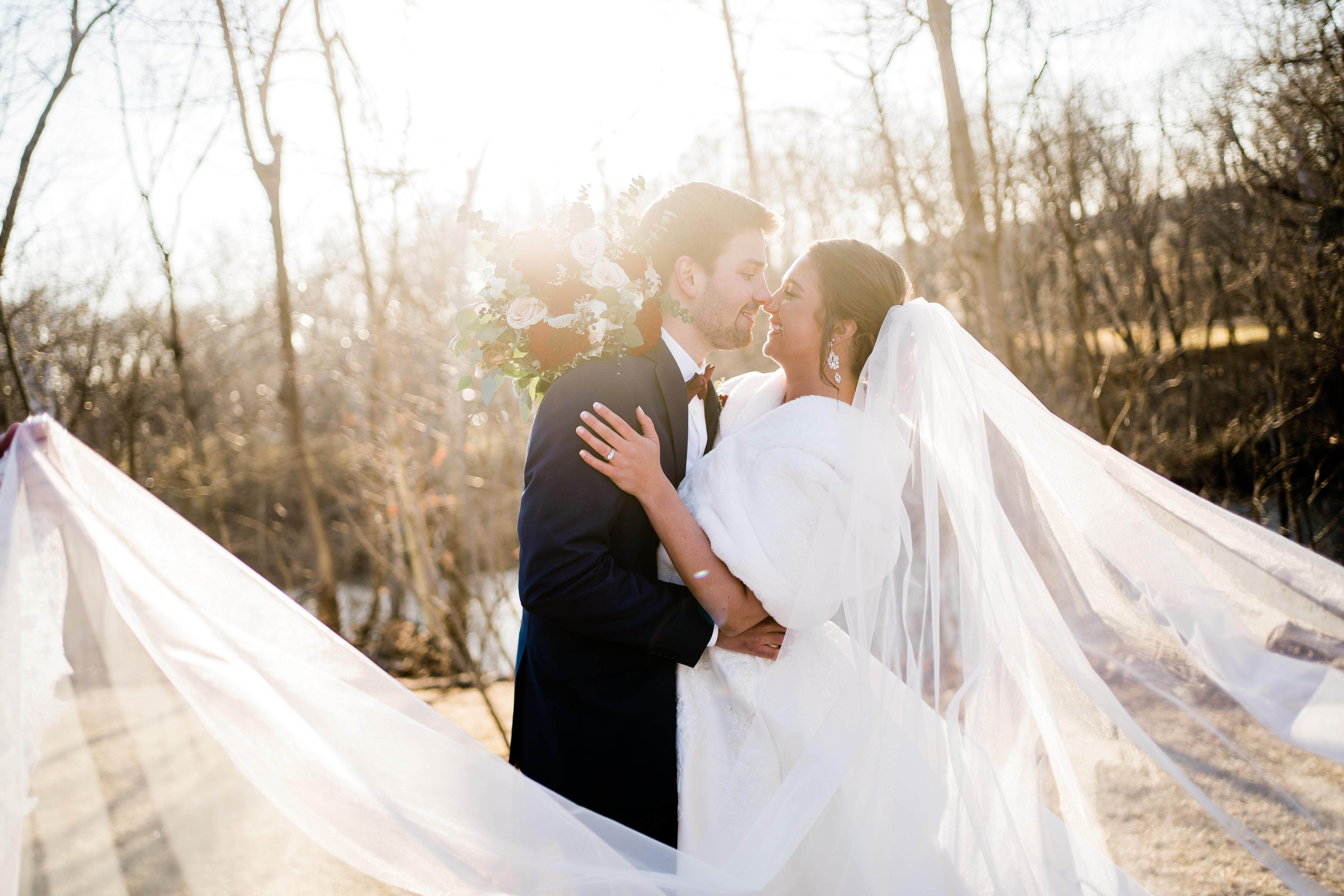 outdoor bridals with bride in a fur coat being dipped by her groom as they kiss and the shine shines through the trees behind them