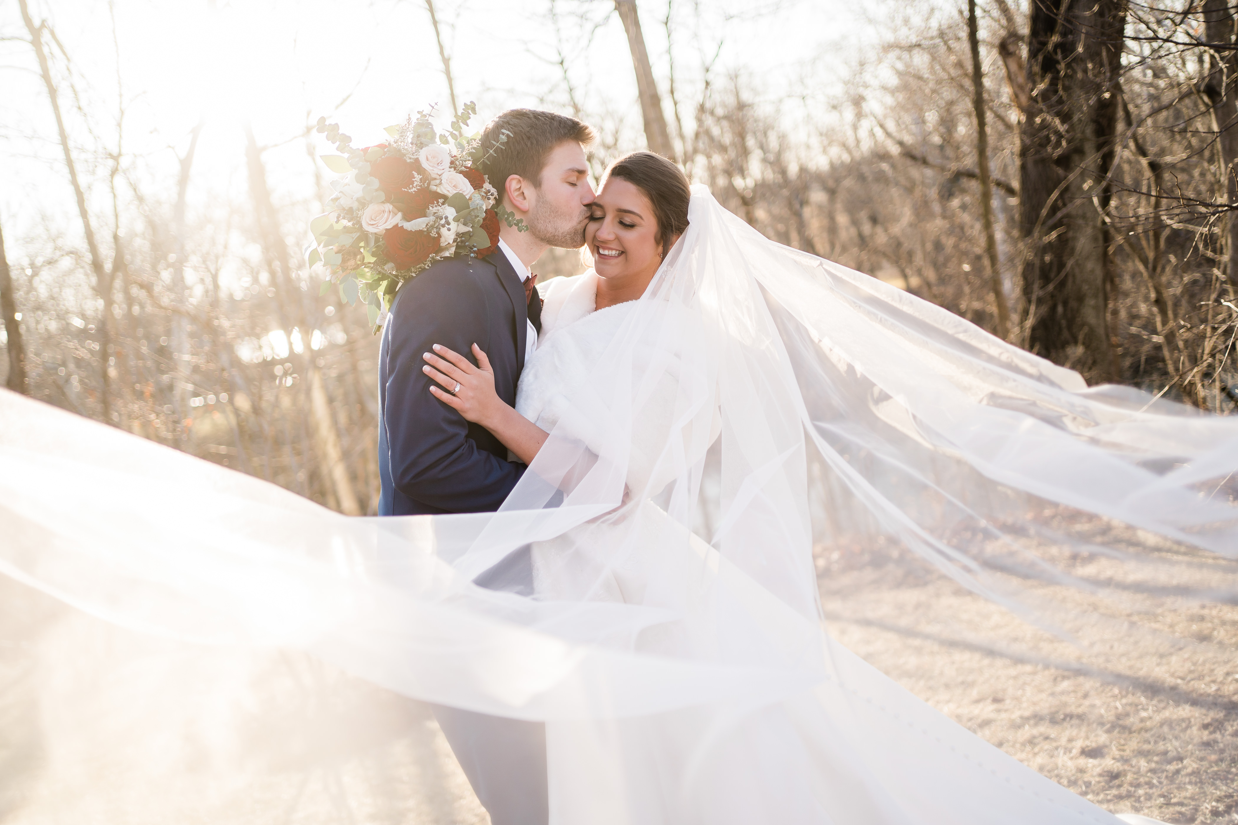 bridal portraits at sunset at The Bluffs at Conner Prairie wedding venue with brides veil in the wind as the groom kisses her cheek