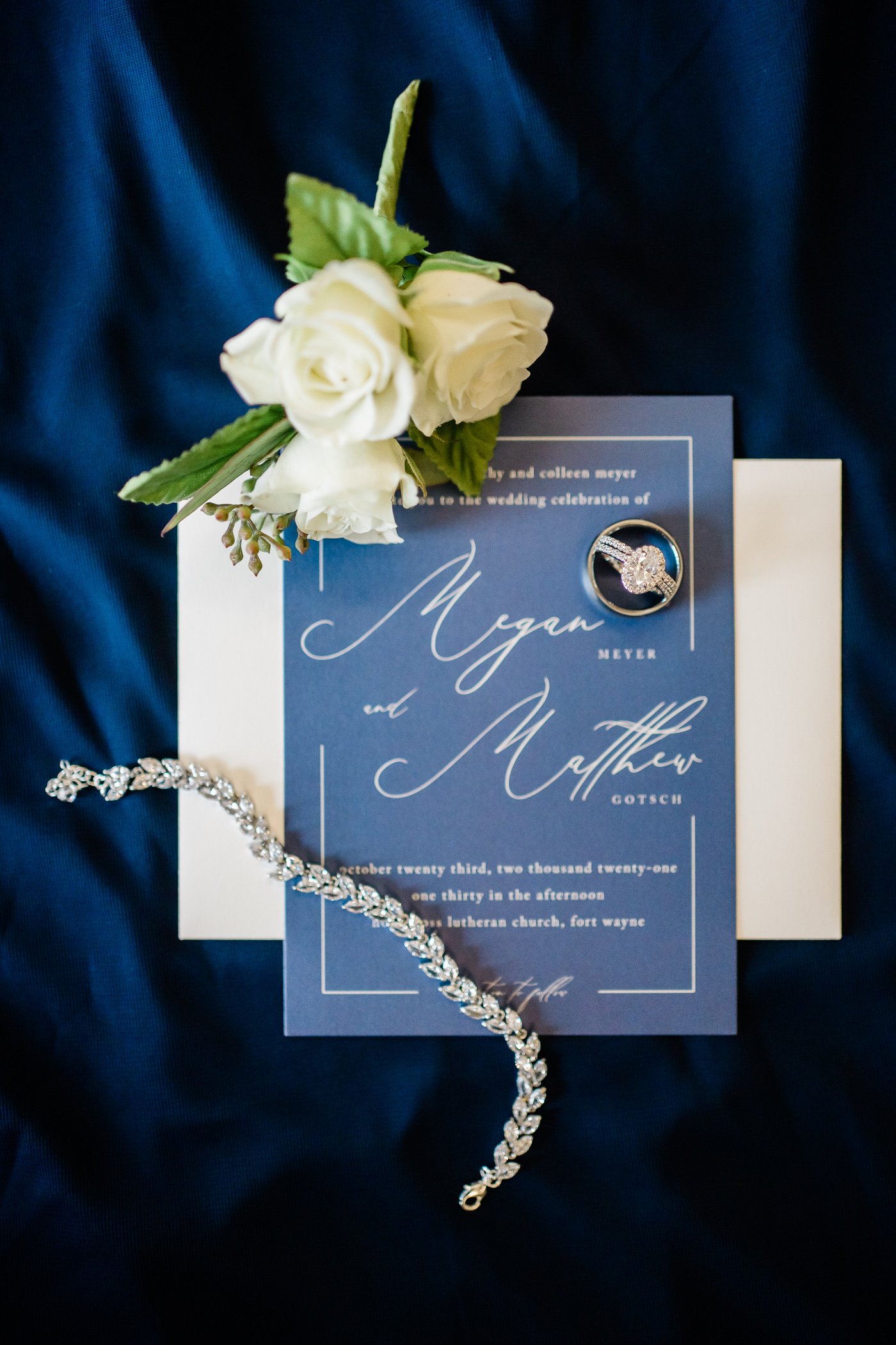 blue wedding invitation with white roses surrounding it as well as the wedding jewelry