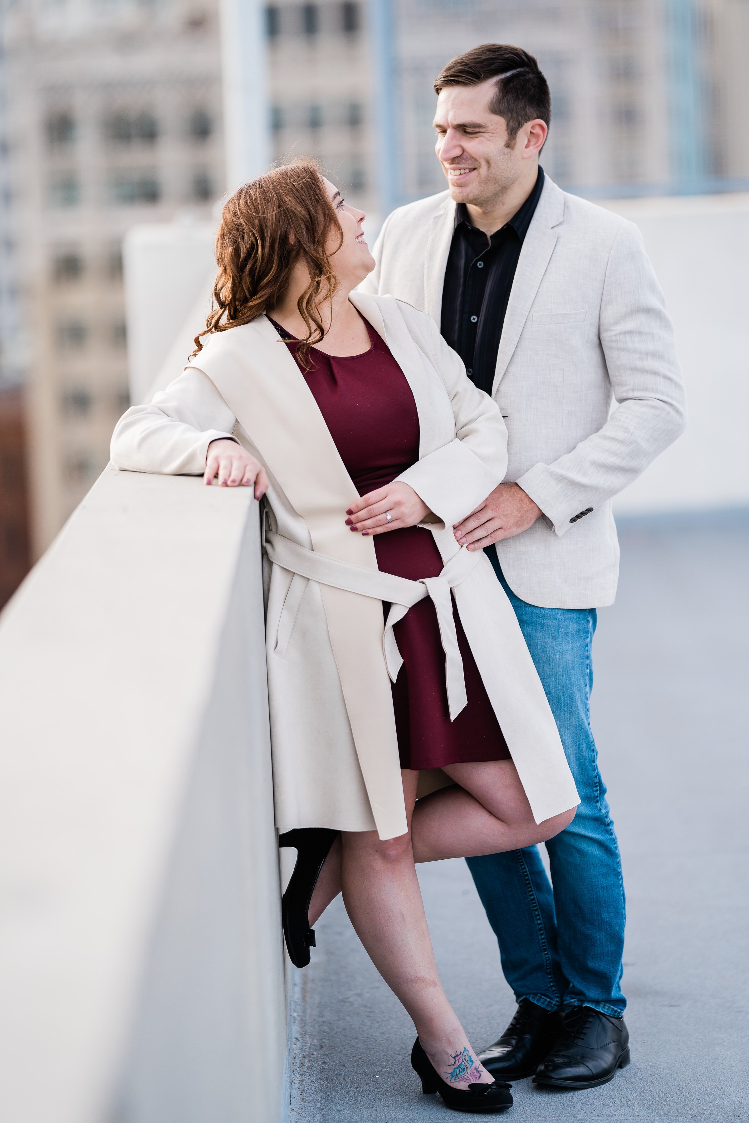 woman leaning against a wall on the top of a parking garage as her fiance leans into her and holds his arm around her