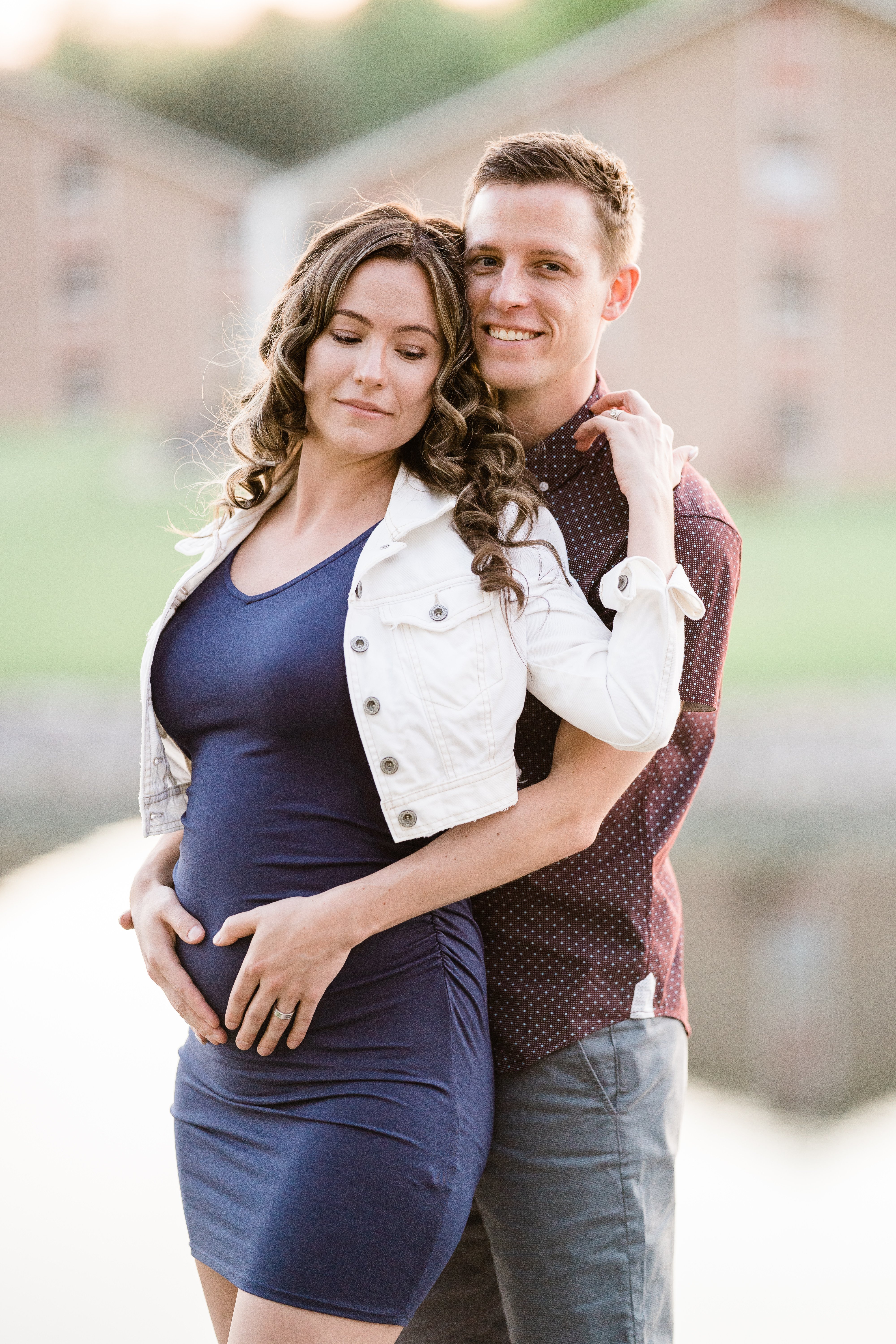 Fort Wayne maternity session with husband standing behind wife and holding her baby bump in front of a lake