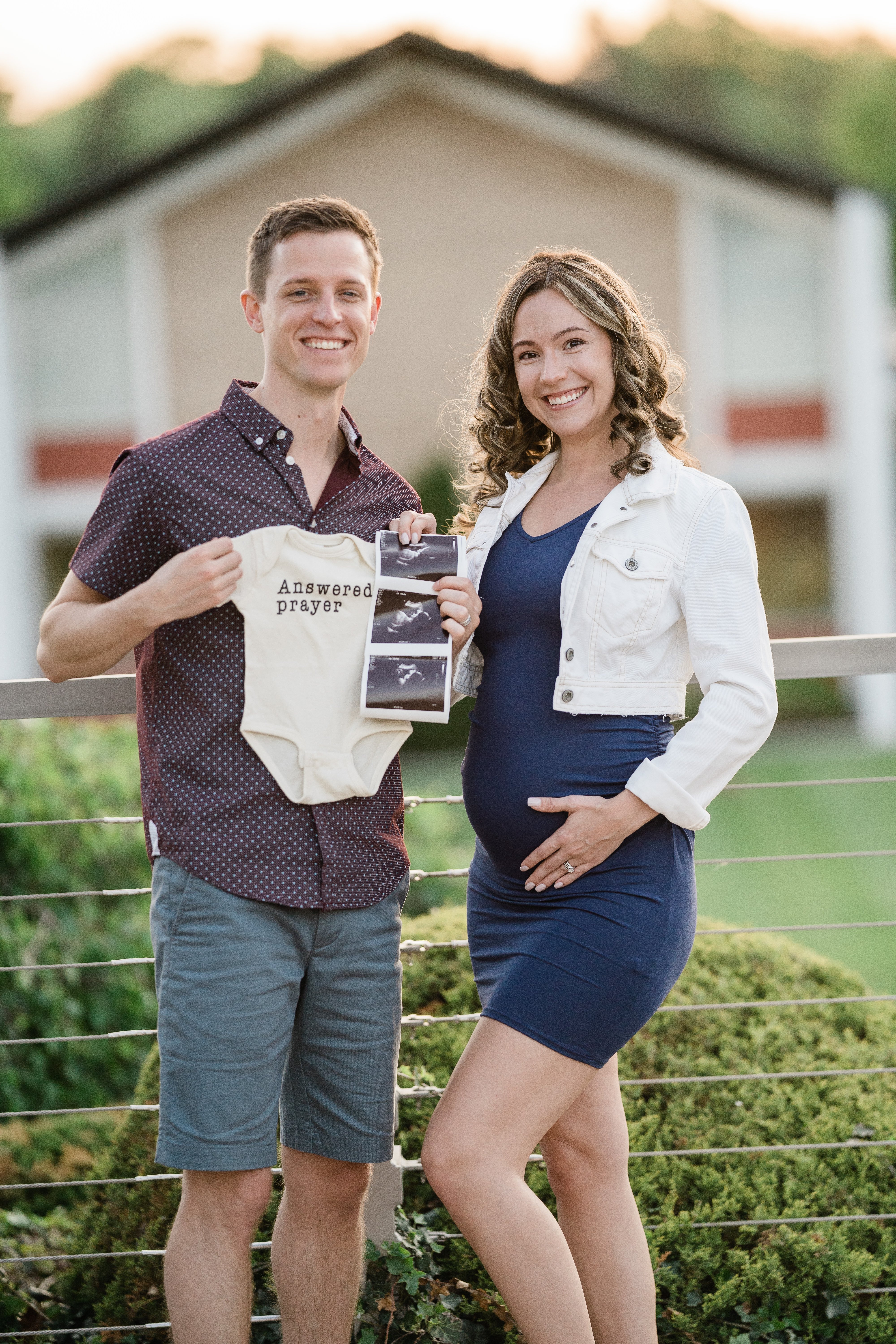 husband and wife standing together for their Fort Wayne maternity session holding up a onesie and ultrasound
