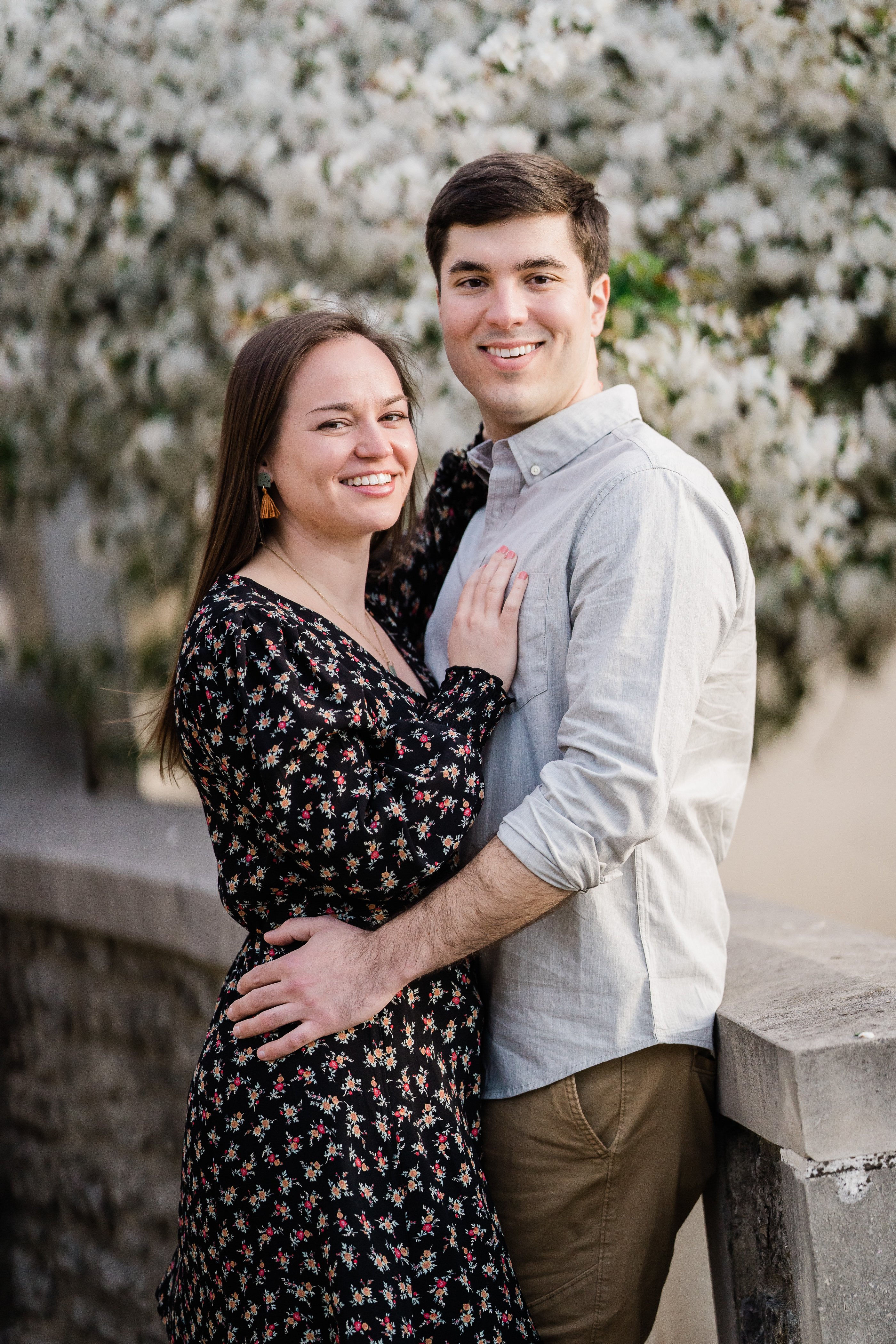 Fort Wayne engagement session in a white blossom orchard with woman in a black dress with small white dots and the man in a light grey button down