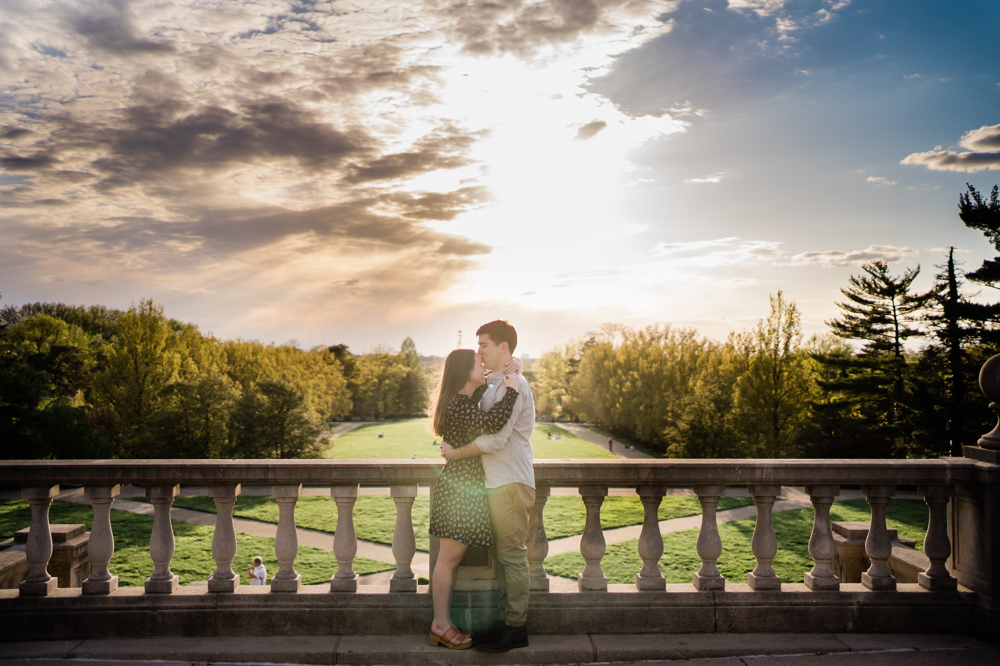 man and woman embracing as the sun sets in the distance over the tree line as they stand on a stone balcony captured by fort wayne wedding photographers