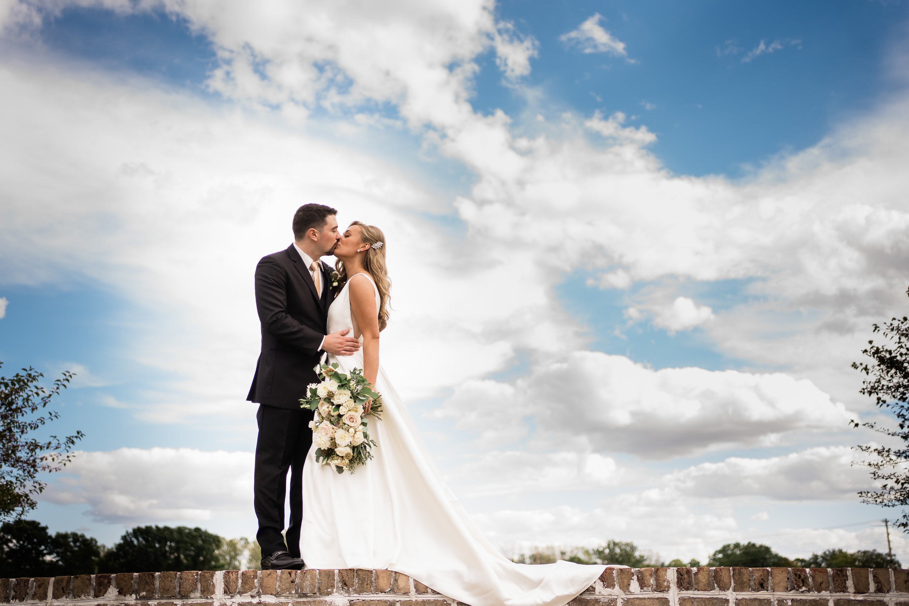 luxury wedding in Fort Wayne with bride and groom standing on a wall in a courtyard and kissing with a blue sky behind them 