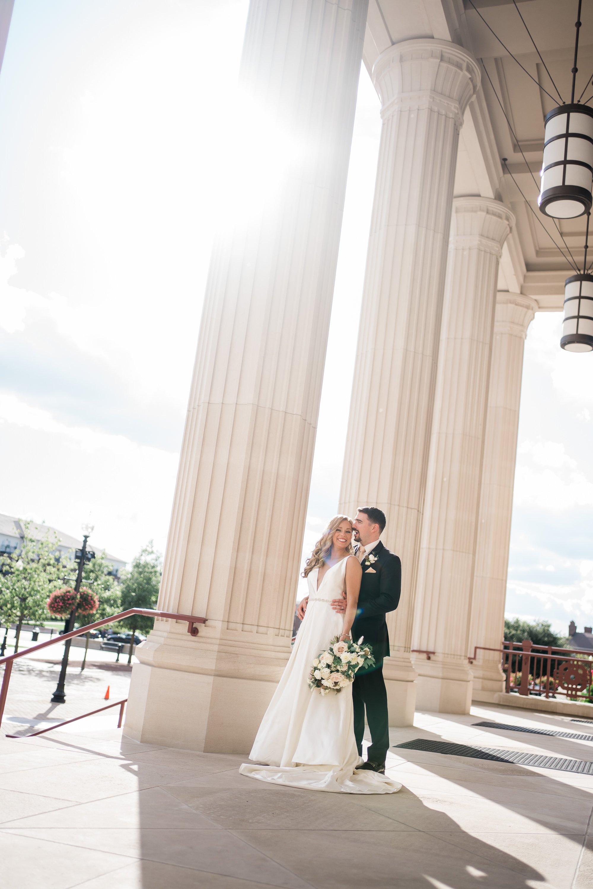 bride and groom embrace next to a tall pillar with the sun shining through onto them