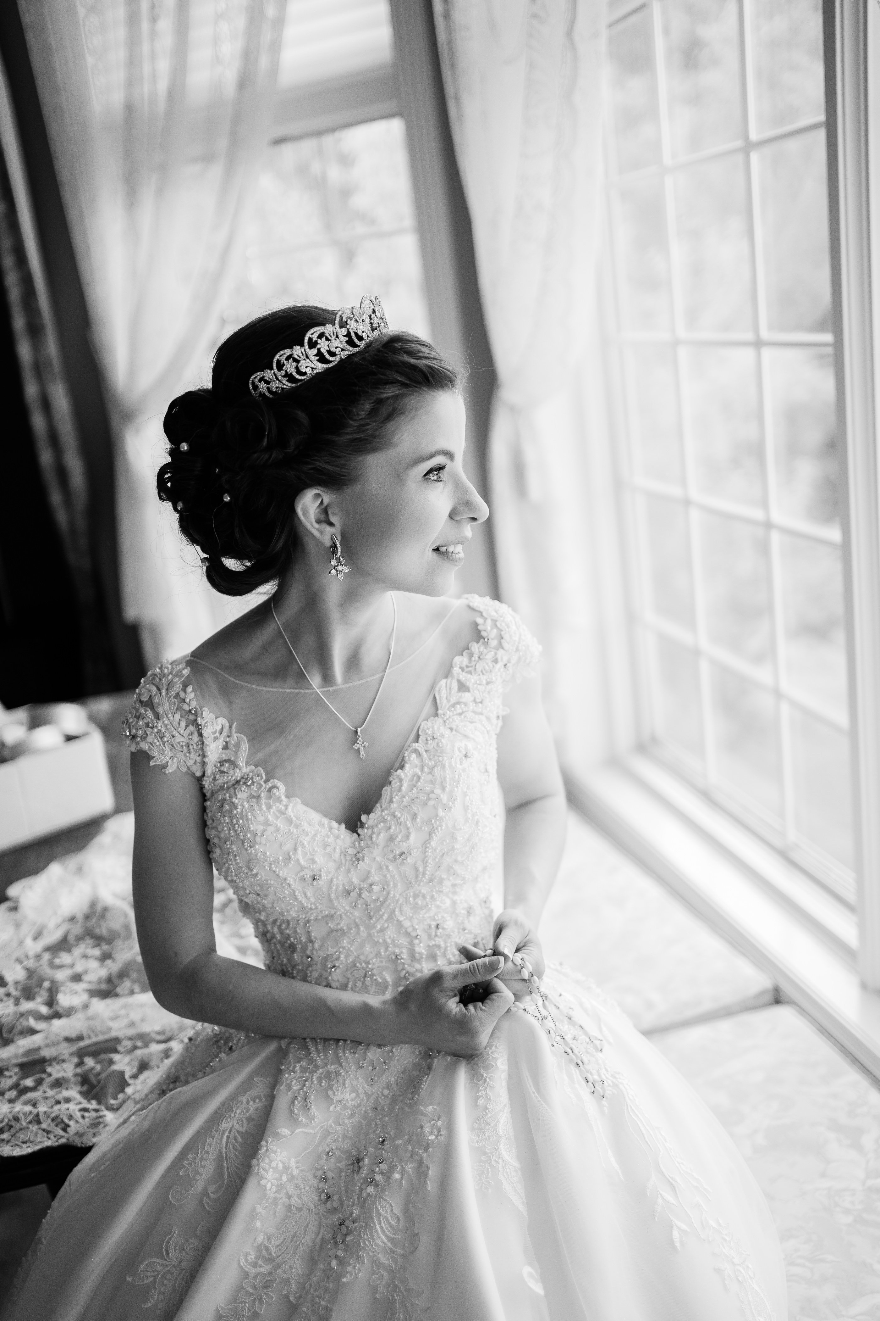 black and white bridal portrait of luxury wedding day as the bride sitting next to a window and looks out as she wear her lace ballgown wedding dress and crown 