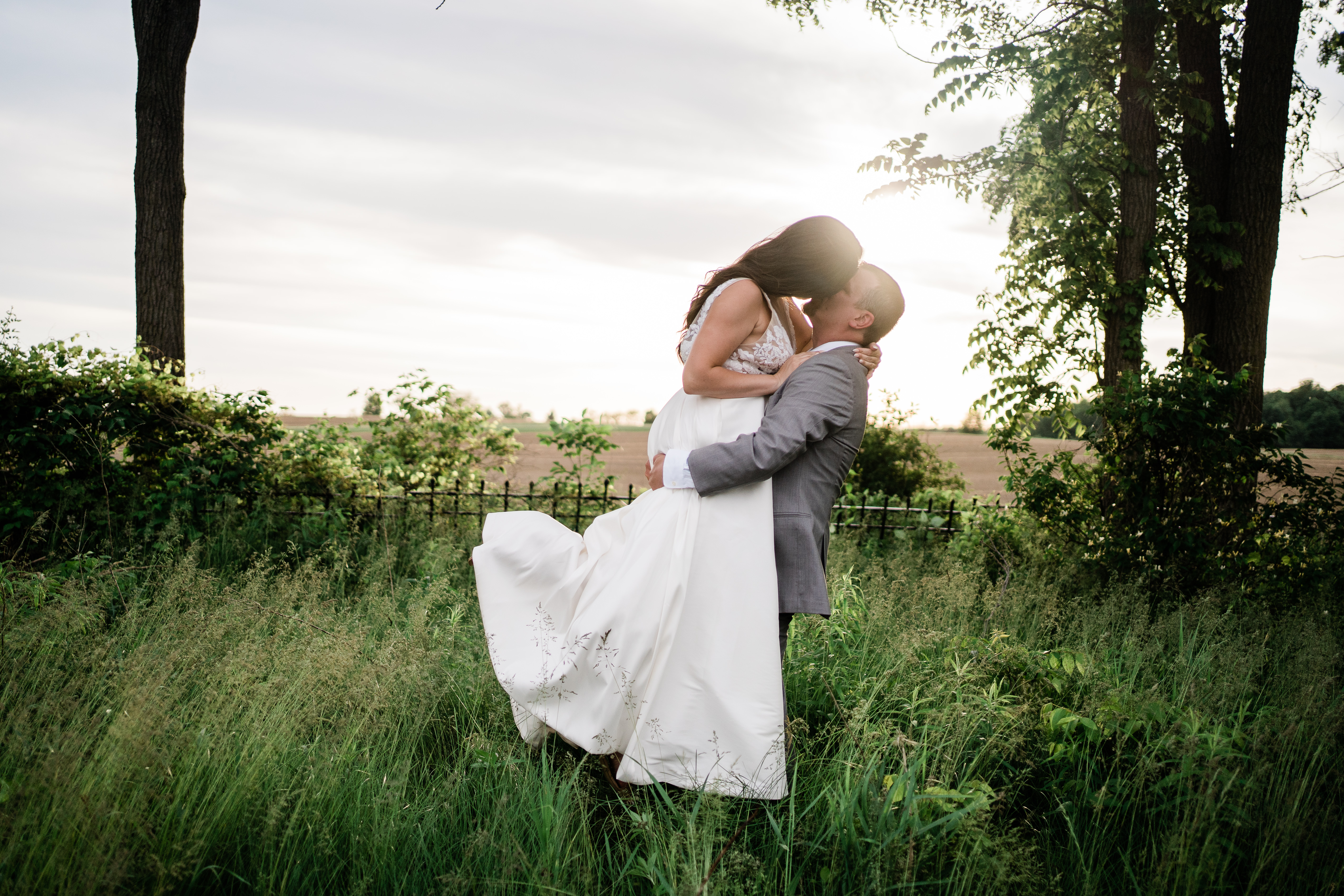 groom lifts up his bride and kisses her in a field in her wedding dress from Fort Wayne Wedding dress shops