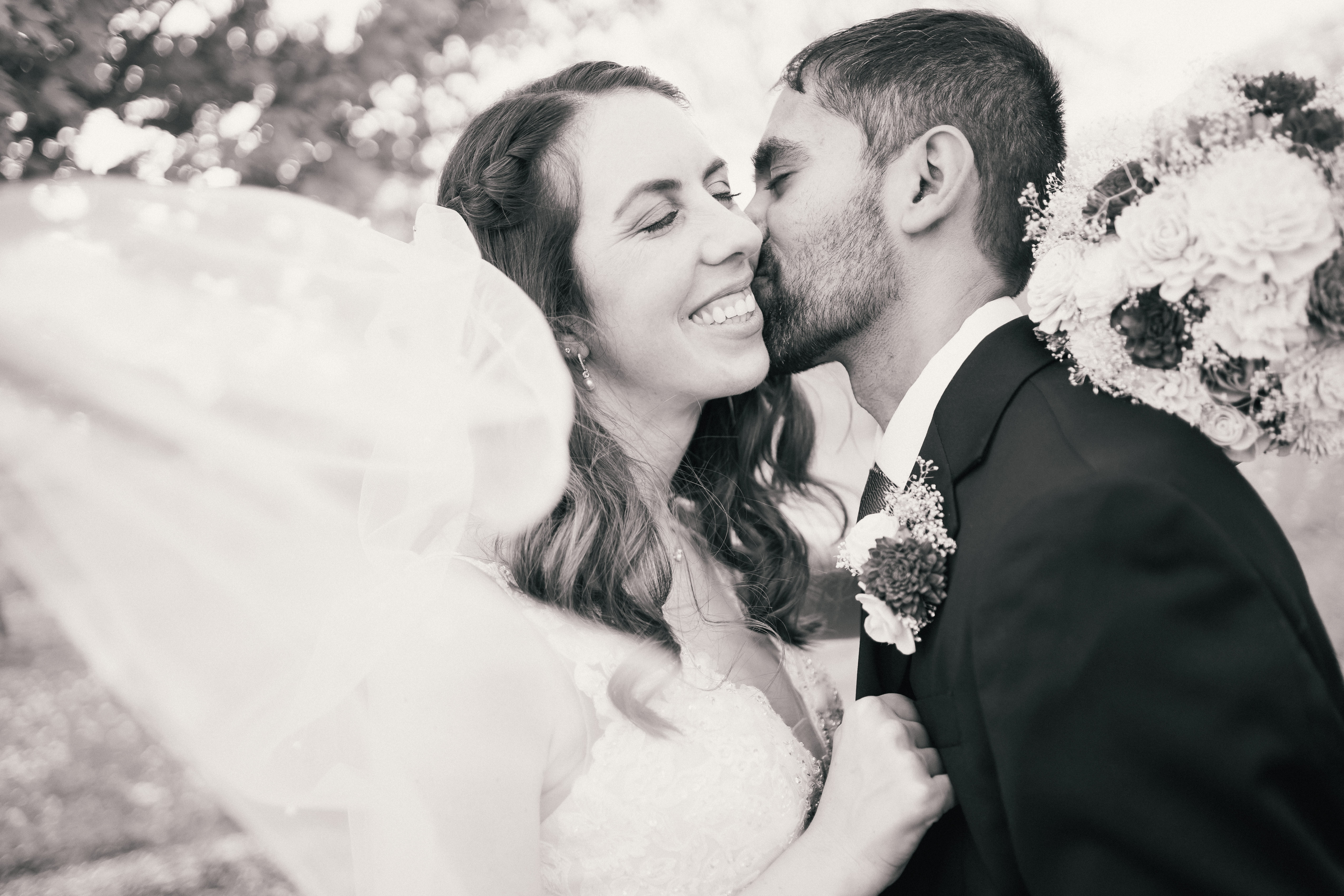 black and white wedding photos outdoors with groom kissing brides neck as she smiles and her veil flows around her