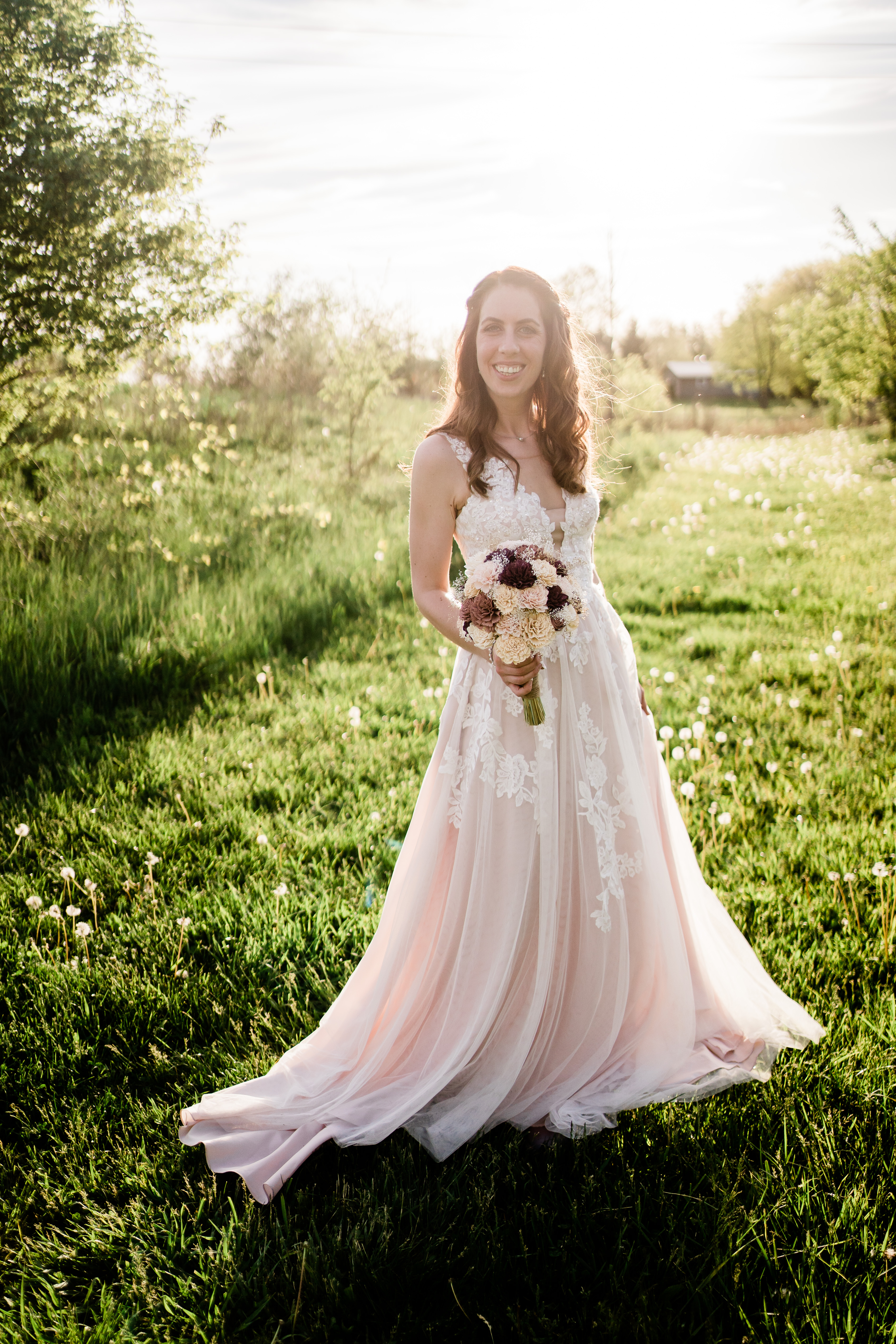 bride standing in a field with the sun setting behind her and holding her bridal bouquet while smiling