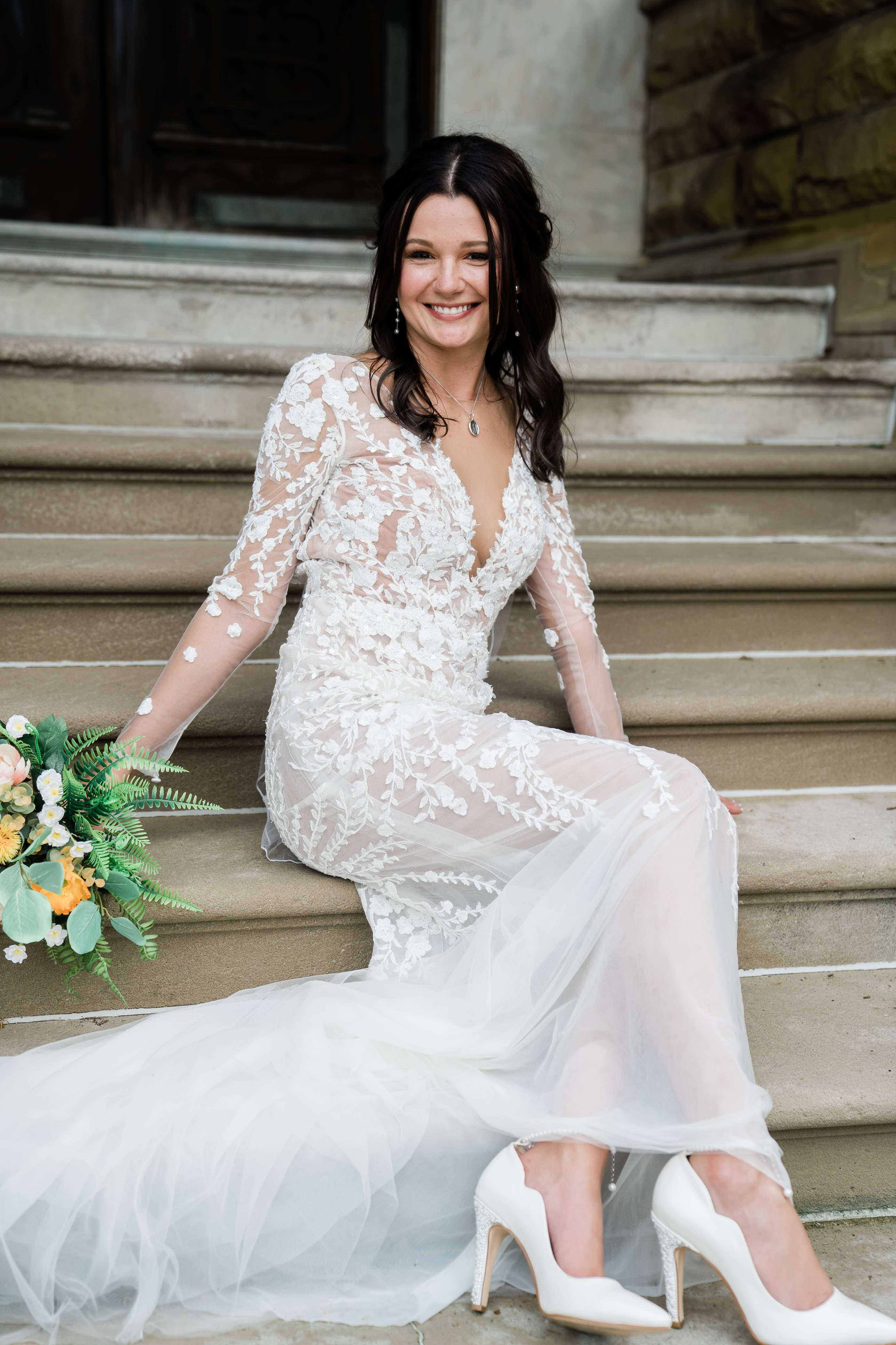 bridal portrait of bride in a floral lace wedding dress with long sleeves sits on outdoor stairs at her wedding chapel