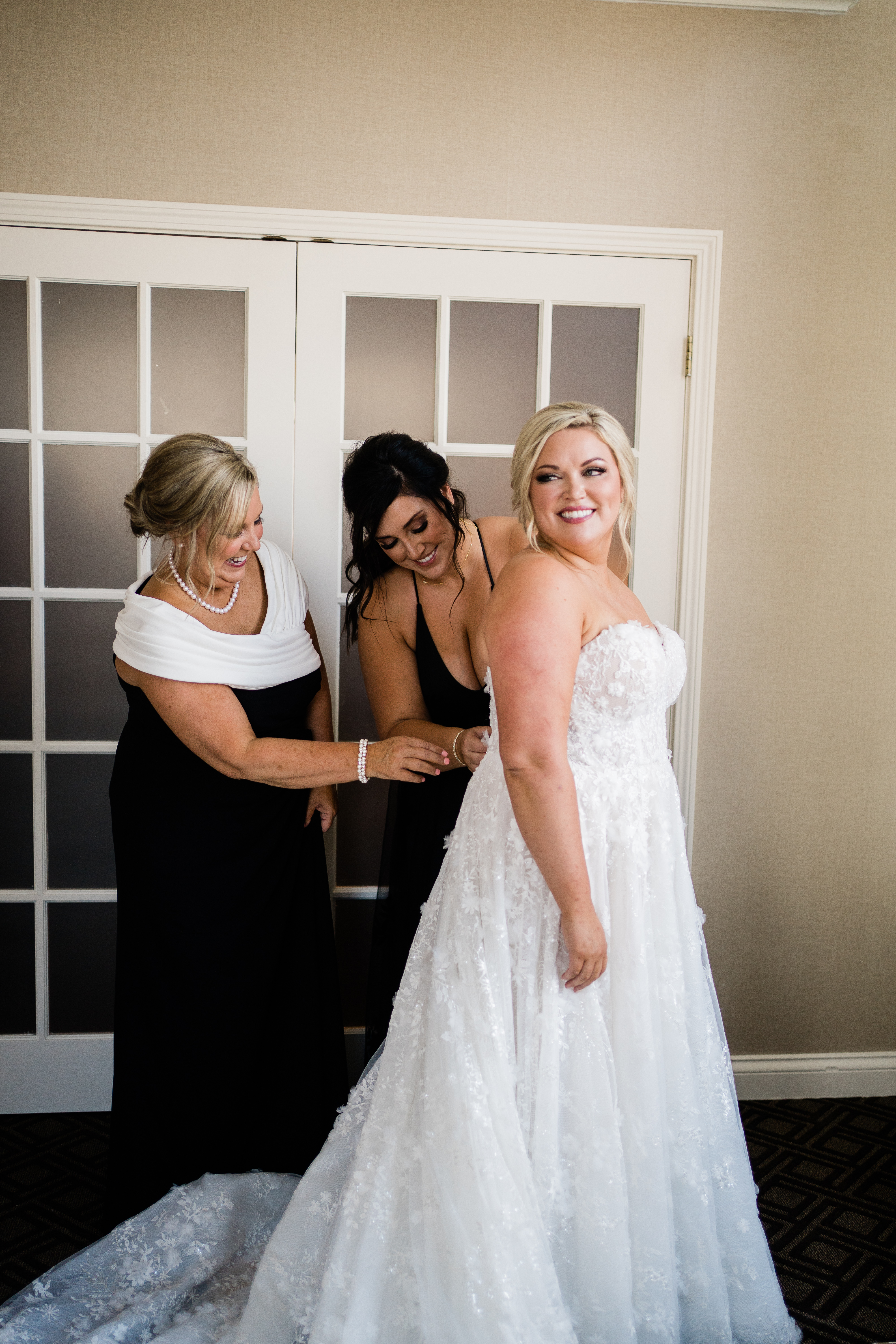 mother of the bride and mother of the groom help zip up the brides dress as she gets ready taken by Fort Wayne wedding photographers